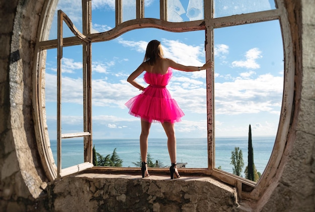 Garota sexy em um lindo vestido rosa no fundo de uma janela velha em um castelo abandonado