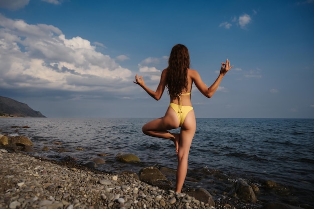 Garota sexy em um maiô com uma figura esbelta fazendo exercícios de ioga na praia à beira-mar
