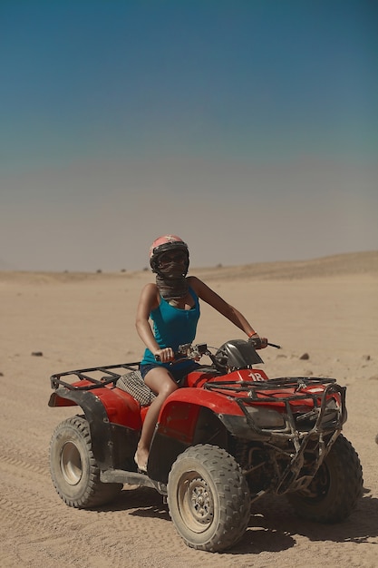 Garota sexy modelo com corpo perfeito e bronzeado, de óculos escuros e capacete, sentada no ATV e posando no deserto. Mulher segurando o volante e posando na estrada no deserto