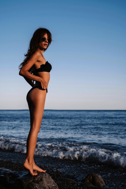Garota sexy morena em um maiô preto com uma figura esbelta posando na praia à beira-mar