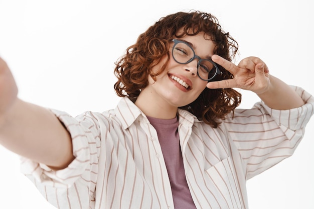 Garota sincera feliz diz peito e tira selfie segura o celular da câmera e posa para foto com gesto de paz vsign sorrindo amplamente em pé sobre fundo branco