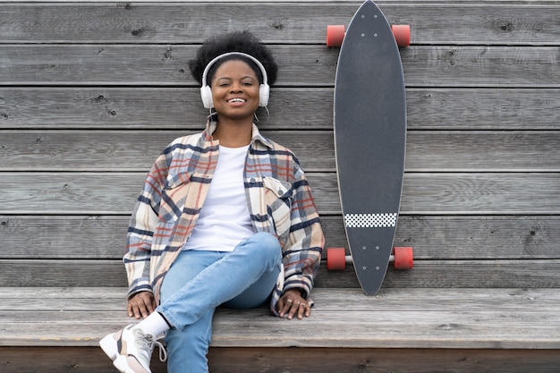 Garota skatista africana feliz com skate ouve música relaxada ao ar livre no parque espacial urbano