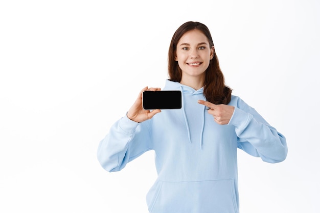 Garota sorridente alegre apontando para smartphone virada horizontalmente mostrando aplicativo ou videogame em pé contra fundo branco