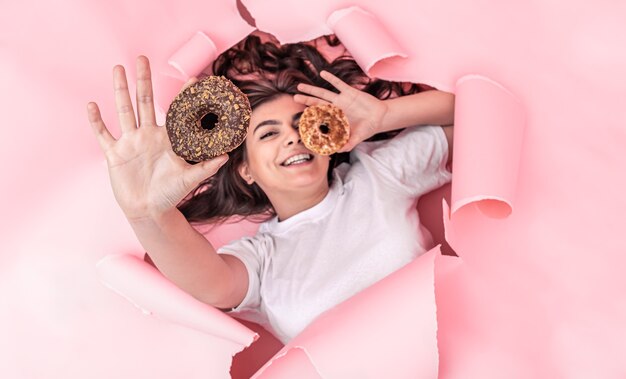 Garota sorridente atraente com rosquinhas apetitosas olha para fora de um buraco em um fundo de papel rosa, vista superior.