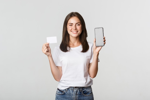 Foto garota sorridente atraente olhando satisfeita e mostrando o cartão de crédito, a tela do telefone móvel.