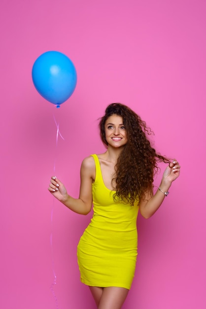 Garota sorridente de vestido amarelo segurando balão de ar azul no fundo rosa