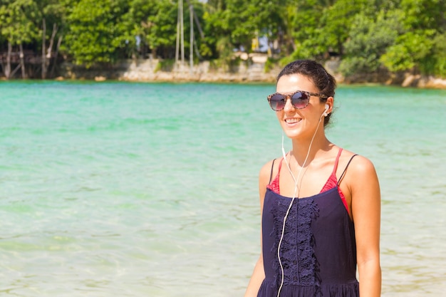 Garota sorridente de vestido azul ouvindo música com fones de ouvido enquanto caminhava na praia na tailândia