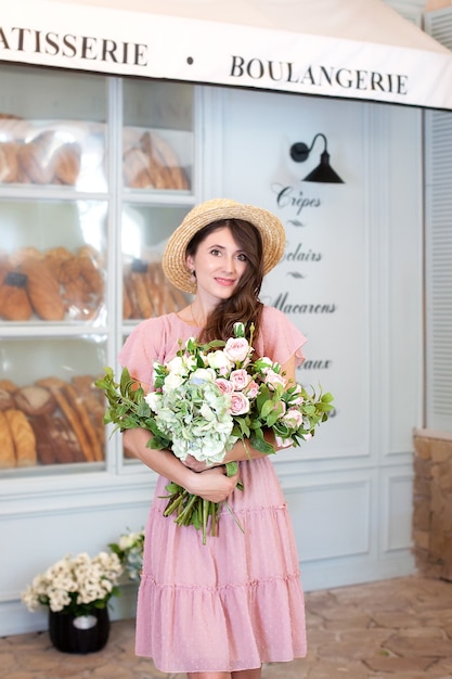 garota sorridente em pé perto da janela da padaria com chapéu de palha e um buquê de flores
