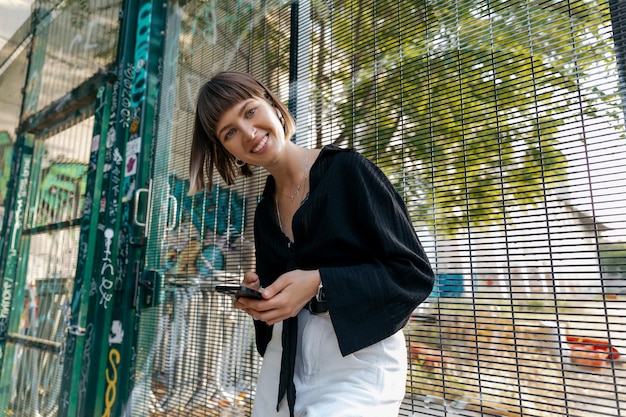 Garota sorridente feliz com emoções verdadeiras posando para a câmera ao ar livre com smartphone Adorável senhora elegante em roupas casuais conversando online com amigos e aproveitando a noite de verão