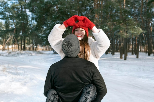 Garota sorridente feliz mantém as mãos em sinal de forma de coração Jovem casal amoroso na floresta de inverno Dia ensolarado e gelado