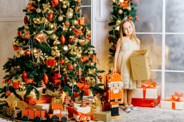 Garota sorridente feliz no vestido festivo em pé perto da árvore de natal com caixas de presente e posando para a câmera