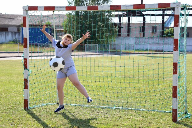 Garota sorridente gordinha em roupas esportivas fica no portão de futebol e quer parar a bola