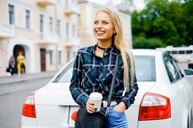 Garota sorridente no centro da cidade com café para viagem perto do carro