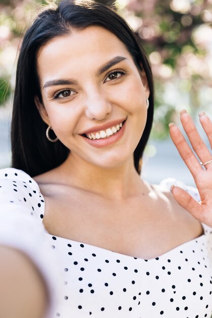 Garota sorridente tendo videochamada em smartphone no parque da cidade amigável mulher acenando com a mão para a câmera