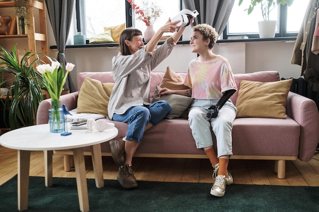 Garota sugerindo sua amiga com braço protético para jogar realidade virtual enquanto eles estão sentados no sofá na sala de estar