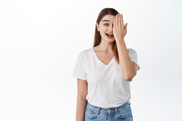 Garota surpresa e feliz cobre metade do rosto sorrindo e ofegando espantado verificando algo incrível em roupas casuais contra fundo branco