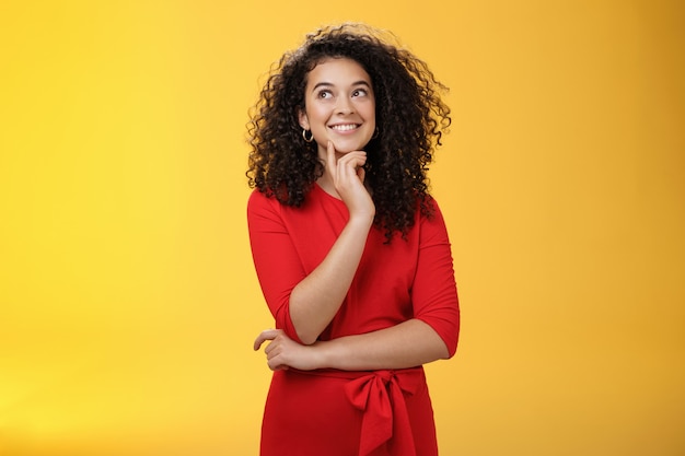 Garota tendo uma ideia interessante, sorrindo, sentindo-se confiante no plano pode funcionar. Mulher feliz encantadora com cabelo encaracolado com vestido vermelho olhando pensativa para o canto superior esquerdo, pensando sobre a parede amarela.