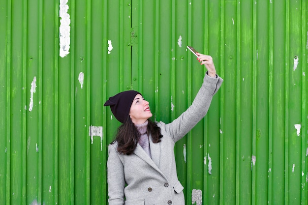 Garota tira uma selfie em uma porta industrial verde. vestindo um chapéu e casaco. lugar para escrever. vestido com um pagode frio. roupas e estilo. emoções naturais