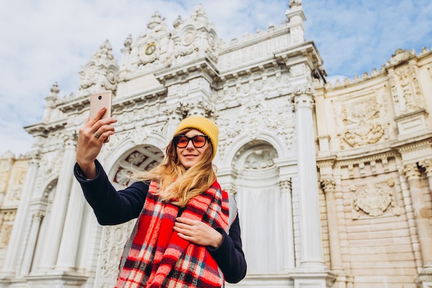 Foto garota toma uma selfie na praça em frente a dolmabahce, istambul, turquia. uma jovem turista de chapéu e casaco é fotografada ao telefone em frente ao palácio do sultão, em istambul.