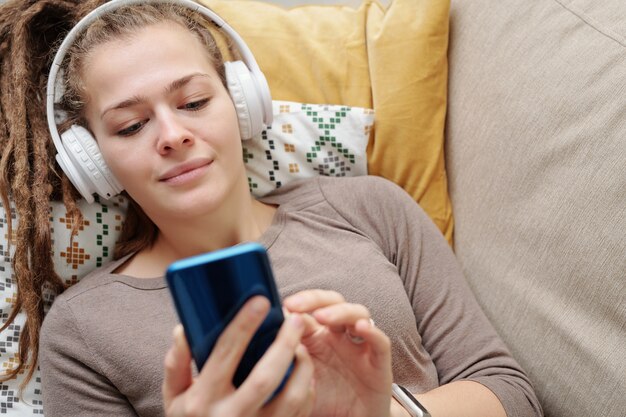 Garota tranquila com dreads ouvindo música em fones de ouvido e rolando no smartphone enquanto relaxa no sofá