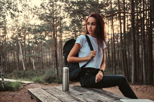 Garota turista sensual sentada em uma mesa de madeira, fazendo uma pausa em uma bela floresta de outono ao pôr do sol.