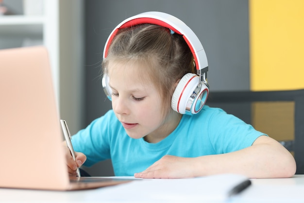 Garota usando fones de ouvido fazendo anotações em um caderno na mesa é um laptop