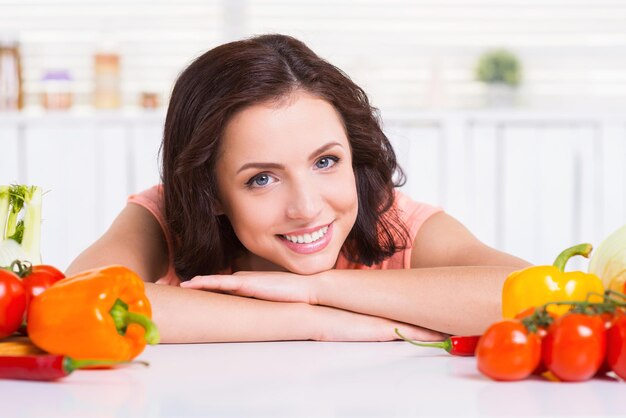 Garota vegetariana. Mulher jovem e atraente encostada na mesa da cozinha e sorrindo enquanto vegetais coloridos perto dela