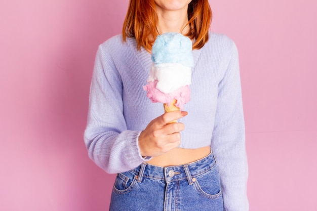 Garota vestindo jeans e suéter azul e segurando um sorvete de algodão doce. Fundo rosa claro.