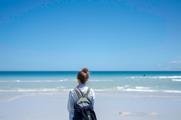 Garota viajando com uma mochila fica em uma praia com areia branca contra um fundo azul-celeste