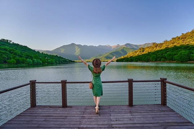 Garota viajante com os braços abertos erguidos, sozinha na borda do cais, olhando para o lago e as montanhas