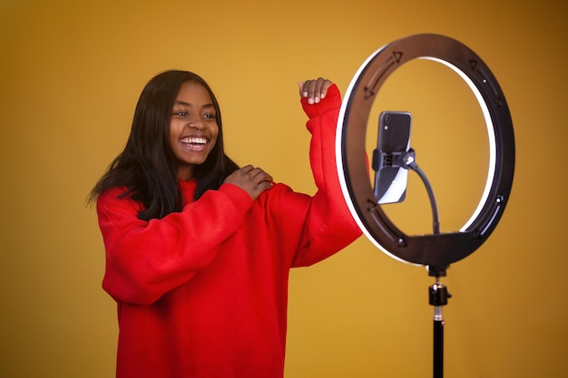 Garota vlogger afro-americana com uma lâmpada de anel iluminada e telefone anunciando um capuz vermelho