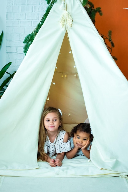 Garotas afro-americanas brancas e negras brincam em uma tenda wigwam crianças riem se divertem infância feliz
