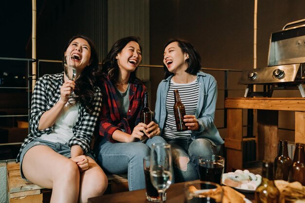 garotas asiáticas se divertindo conversando na festa de churrasco no telhado à noite. jovens senhoras sentadas relaxam nos feriados bebendo álcool cerveja champanhe e vinho. amigos rindo falando de coisas engraçadas.