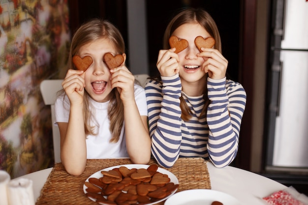 Garotas engraçadas e rindo seguram biscoitos em forma de coração, fecham os olhos e brincam