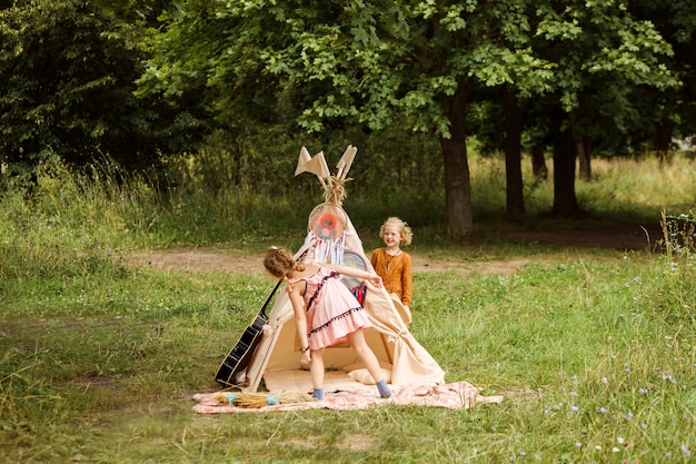 Garotas engraçadas estão brincando ao lado da cabana. as crianças se vestem no estilo boho ou hippie e brincam de esconde-esconde. no verão, ao ar livre na floresta ou no parque. fim de semana em família.