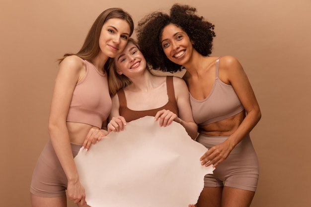 Foto garotas interraciais atraentes em lingerie olhando para a câmera e segurando um cartaz em branco sobre fundo bege