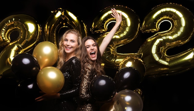 Garotas lindas e felizes em vestidos de festa sensuais elegantes segurando balões de ouro 2022, se divertindo na festa de Natal ou Ano Novo.