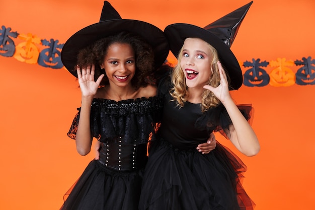 Foto garotas multinacionais encantadas em fantasias pretas de halloween se abraçando e ouvindo isoladas sobre uma parede de abóbora laranja