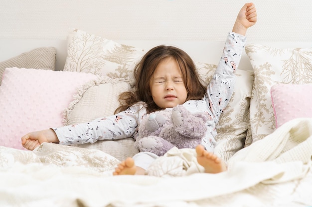 Garotinha adorável e sonhadora se esticando levantando os braços e fechando os olhos com um ursinho de pelúcia na cama macia