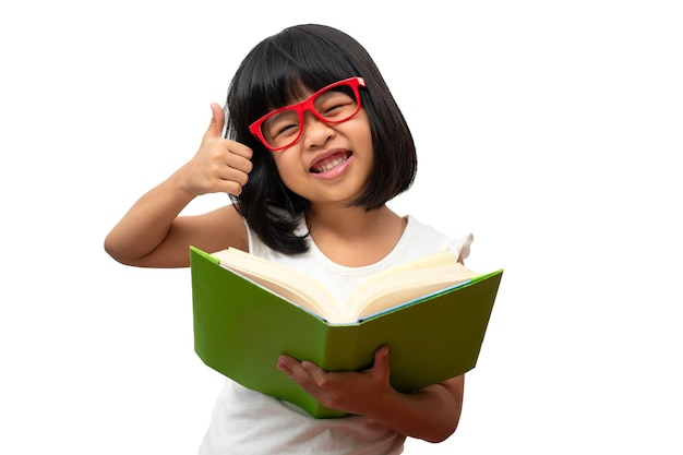 Foto garotinha asiática feliz em idade pré-escolar usando óculos vermelhos, segurando um livro verde e o polegar para cima em branco