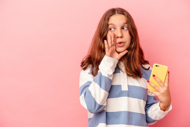 Garotinha caucasiana segurando um celular isolado em um fundo rosa está contando uma notícia secreta de travagem e olhando para o lado