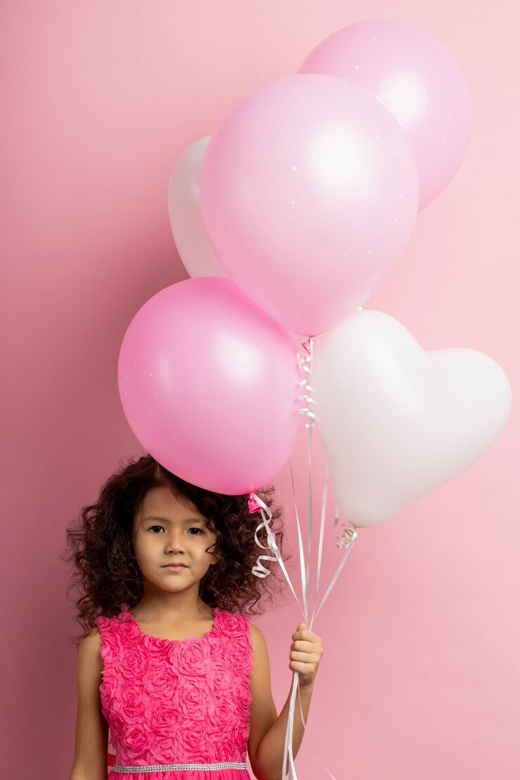 Foto garotinha confusa com cabelos cacheados, usando um vestido lindo, segurando balões de ar branco e rosa, olhando com expressão envergonhada