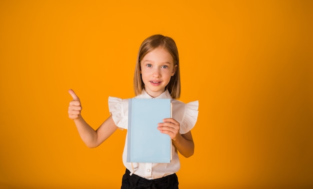 Garotinha de blusa branca segura um caderno azul e mostra a turma com a mão sobre um fundo amarelo com uma cópia do espaço