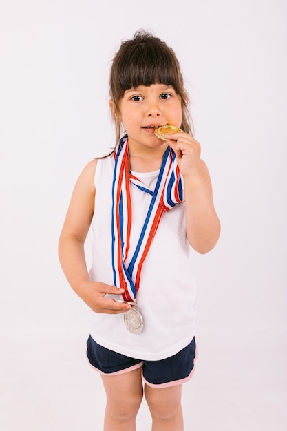 Foto garotinha de cabelos castanhos com medalhas de campeã esportiva, mordendo-as. conceito de esporte e vitória