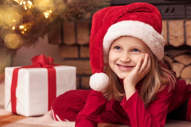 Garotinha de Natal com chapéu de Papai Noel vermelho sonhando perto de árvore de Natal e lareira