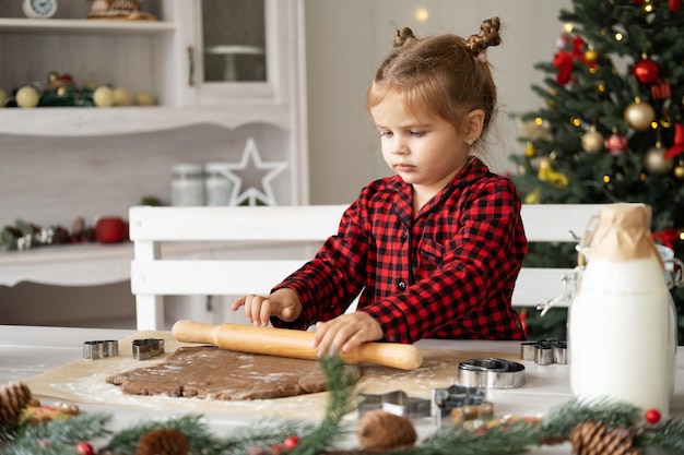 garotinha de pijama vermelho cozinhando pão de gengibre festivo na cozinha decorada de natal