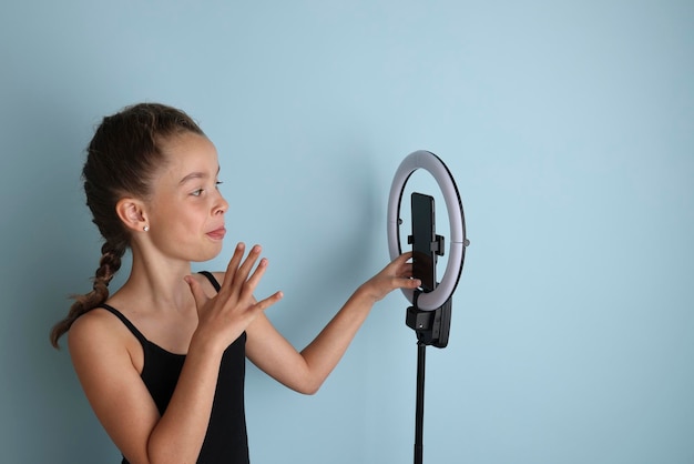 Garotinha emocional em uma camiseta preta em um fundo azul isolado Retrato de estúdio de uma jovem adolescente sorridente vlogger com uma lâmpada de flash de anel e um smartphone Garota tira uma selfie