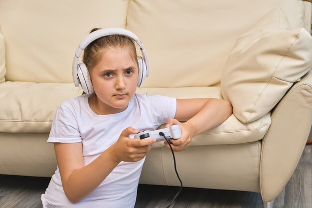 Foto garotinha estressante com fones de ouvido está jogando videogame, olhando para a tela e sorrindo enquanto está sentada no sofá em casa