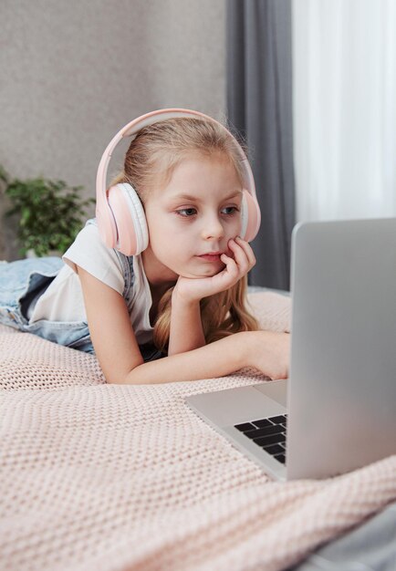 Garotinha feliz sorridente usando laptop em fones de ouvido sem fio na cama em casa