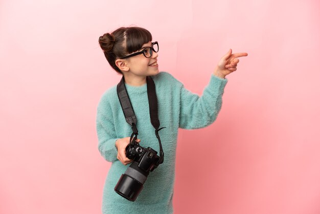 Garotinha fotógrafa isolada em uma parede rosa apontando o dedo para o lado e apresentando um produto